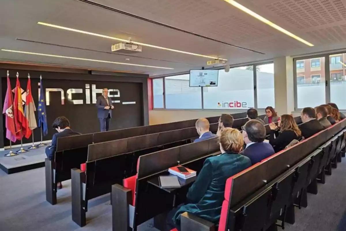 Una persona presenta en un auditorio del INCIBE con varias personas sentadas escuchando y banderas a un lado. Una persona presenta en un auditorio del INCIBE con varias personas sentadas escuchando y banderas a un lado.