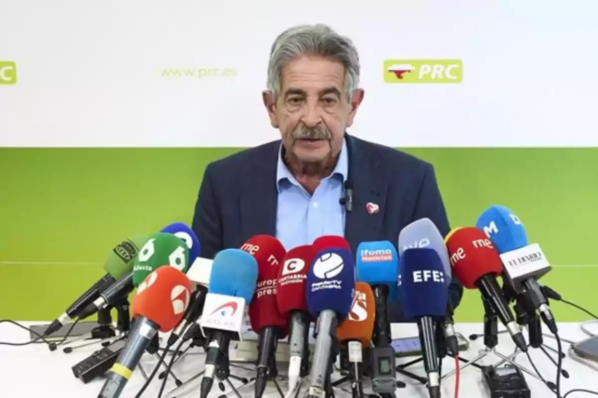 Un hombre de cabello canoso y bigote está hablando en una conferencia de prensa frente a múltiples micrófonos de diferentes medios de comunicación, con un fondo verde y blanco que muestra el logo y sitio web del PRC. Un hombre de cabello canoso y bigote está hablando en una conferencia de prensa frente a múltiples micrófonos de diferentes medios de comunicación, con un fondo verde y blanco que muestra el logo y sitio web del PRC.