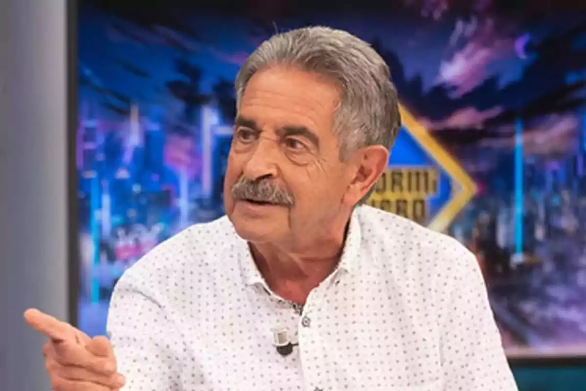 Un hombre de cabello canoso y bigote está hablando en un programa de televisión con un fondo colorido.