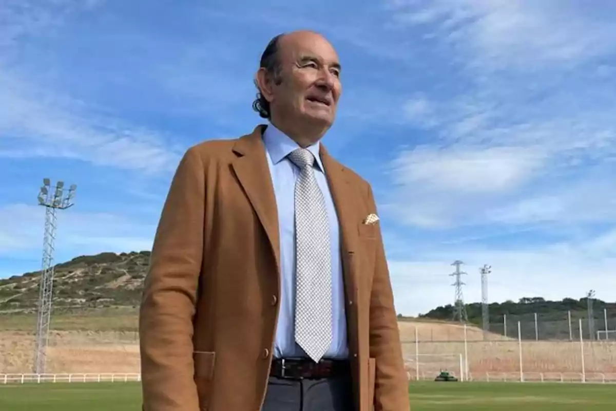 Hombre con chaqueta marrón y corbata de pie en un campo al aire libre con cielo azul y algunas nubes.