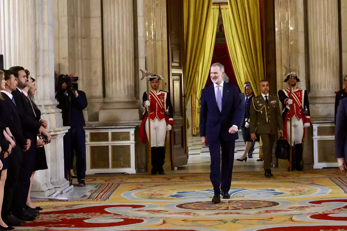 Un hombre con traje oscuro camina por una sala elegante con alfombra decorada y cortinas doradas mientras varias personas lo observan y guardias con uniformes formales están de pie junto a la puerta. Un hombre con traje oscuro camina por una sala elegante con alfombra decorada y cortinas doradas mientras varias personas lo observan y guardias con uniformes formales están de pie junto a la puerta.