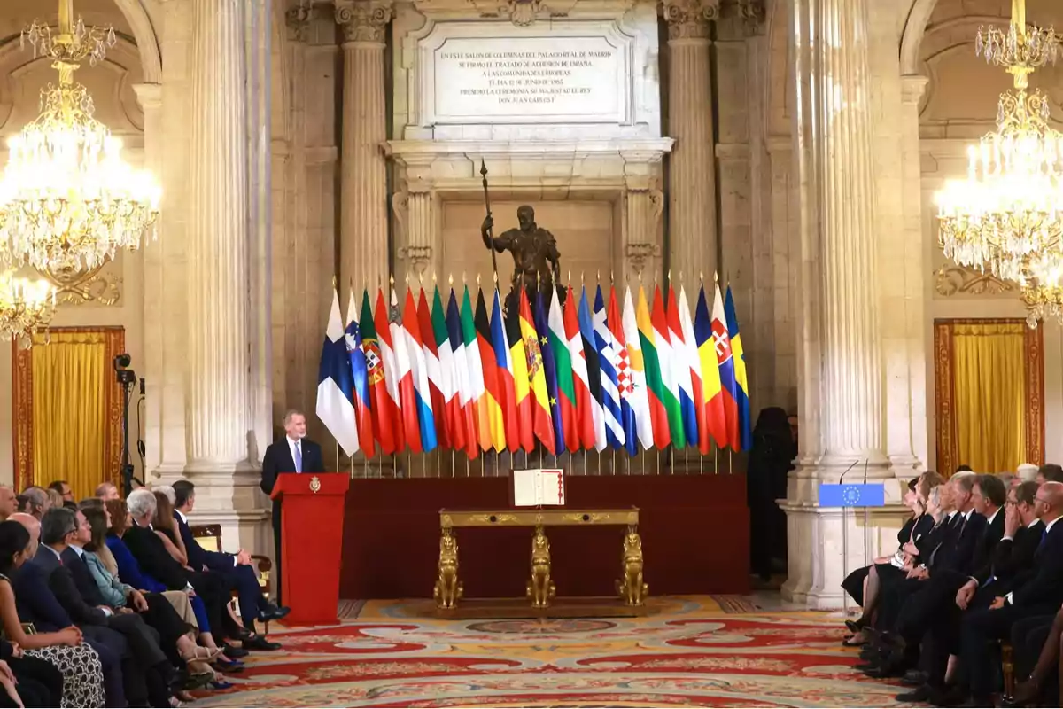 Un hombre da un discurso en un salón elegante con varias personas sentadas y una fila de banderas de diferentes países detrás de él