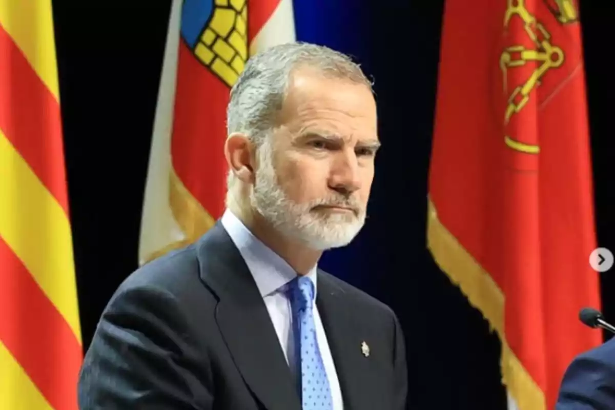 Un hombre con barba y traje oscuro está frente a varias banderas coloridas. Un hombre con barba y traje oscuro está frente a varias banderas coloridas.