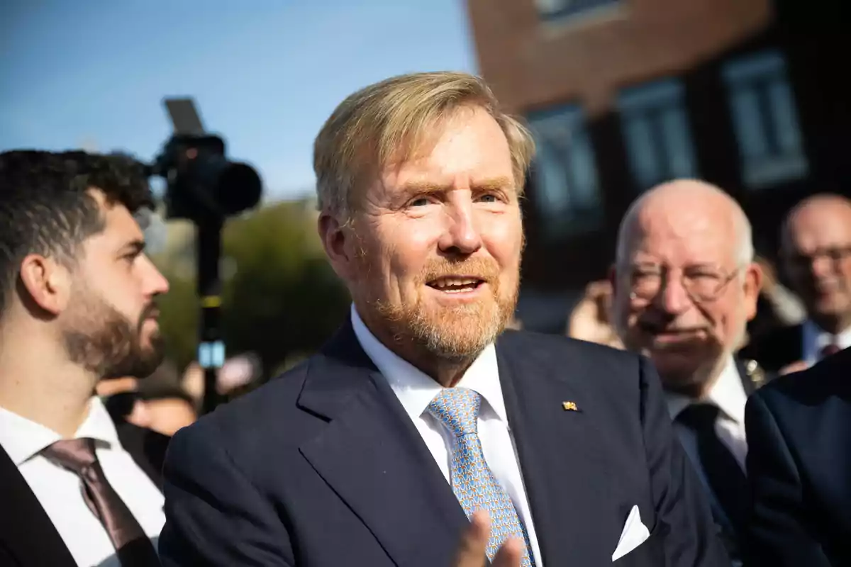 El rey Guillermo de Holanda con traje y corbata sonríe mientras está rodeado de otras personas en un evento al aire libre. El rey Guillermo de Holanda con traje y corbata sonríe mientras está rodeado de otras personas en un evento al aire libre.