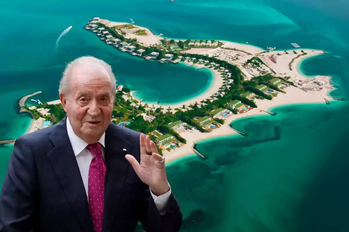 Un hombre mayor en traje oscuro y corbata rosa levanta la mano frente a una isla con playas y edificios rodeados de agua turquesa.