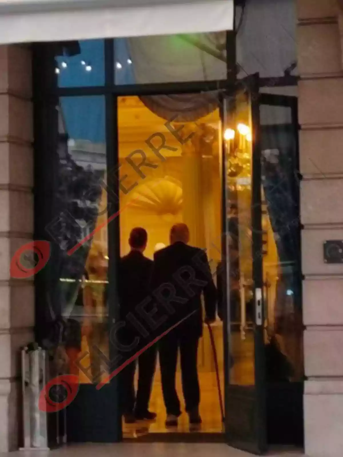 Dos personas de pie en la entrada de un edificio elegante, vistas desde afuera a través de una puerta de vidrio.