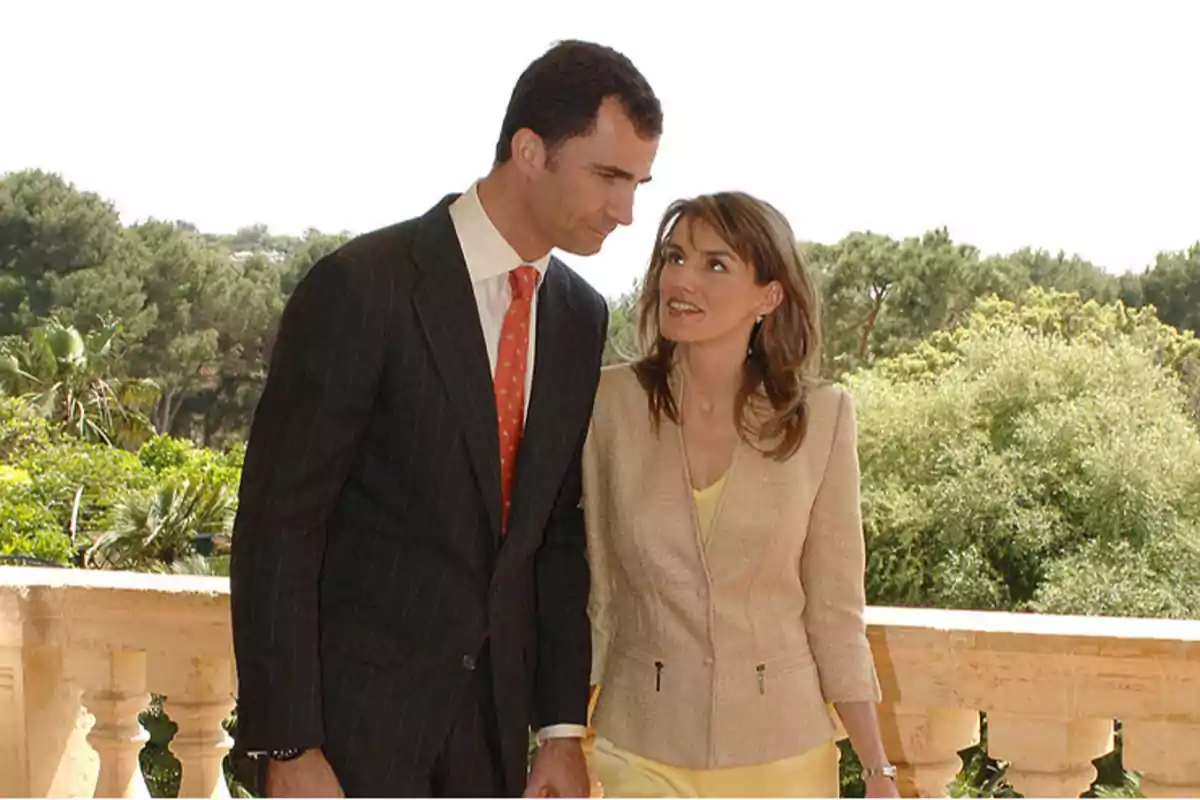 Una pareja vestida formalmente, él con traje oscuro y corbata roja, ella con chaqueta beige y blusa amarilla, de pie en un balcón con un paisaje verde de fondo.