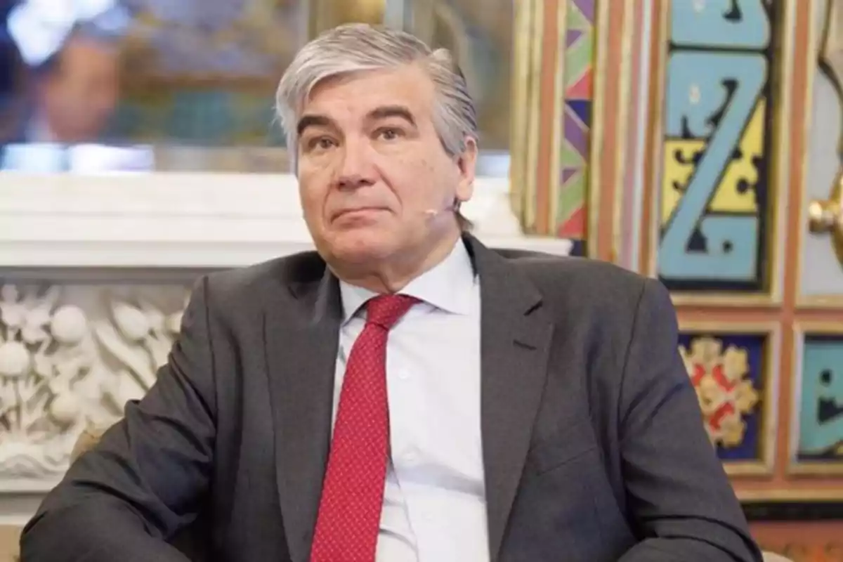Un hombre con traje gris y corbata roja sentado frente a un fondo decorativo. Un hombre con traje gris y corbata roja sentado frente a un fondo decorativo.