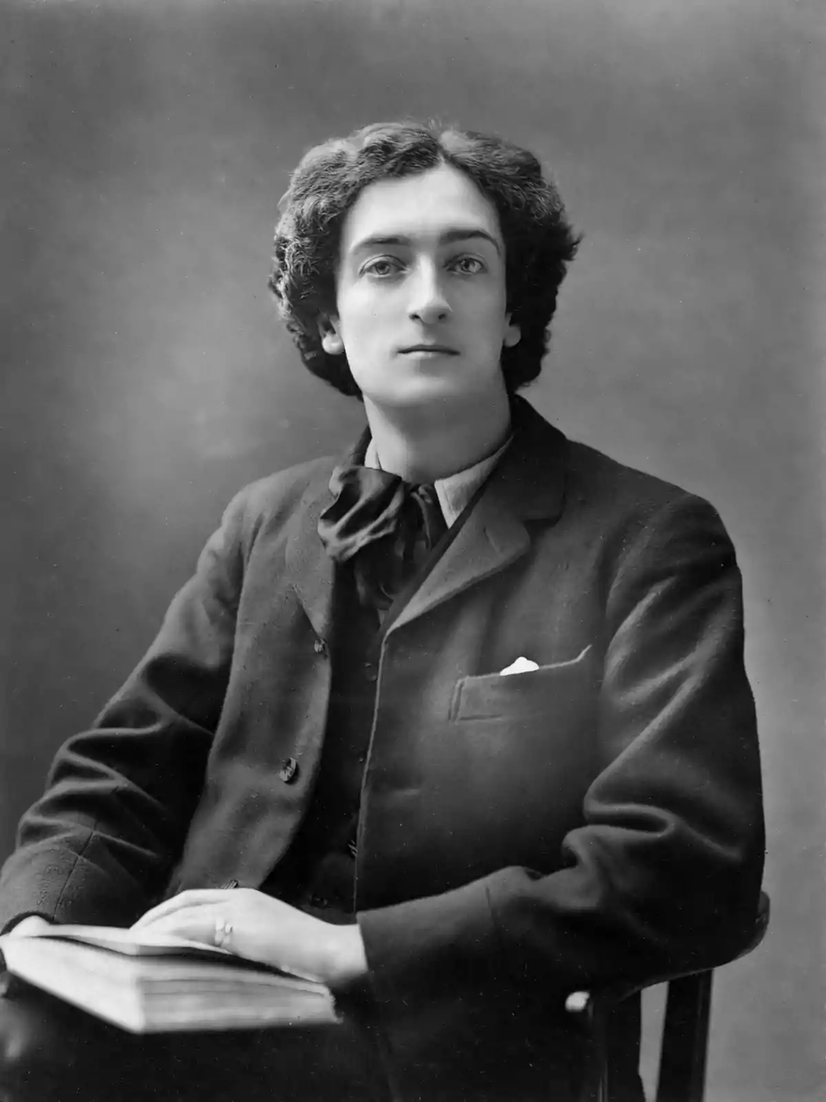 Un hombre joven con cabello rizado y traje oscuro está sentado, mirando hacia la cámara, con un libro en sus manos. Un hombre joven con cabello rizado y traje oscuro está sentado, mirando hacia la cámara, con un libro en sus manos.