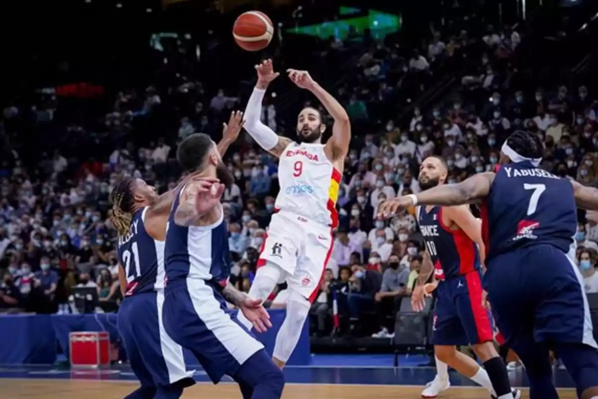 Jugador de baloncesto de España lanza el balón mientras es defendido por varios jugadores de Francia en un partido con público en las gradas Jugador de baloncesto de España lanza el balón mientras es defendido por varios jugadores de Francia en un partido con público en las gradas