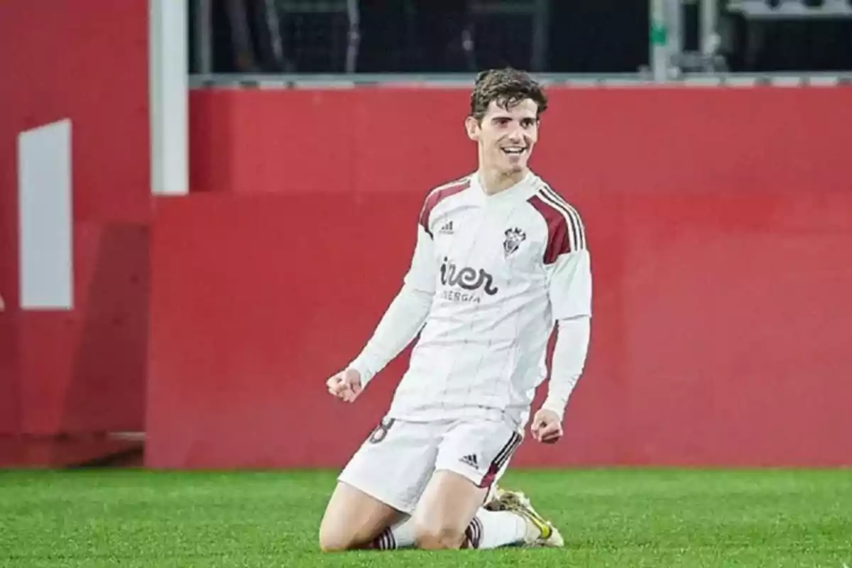 Jugador de fútbol celebrando de rodillas en el campo con uniforme blanco y detalles en rojo.