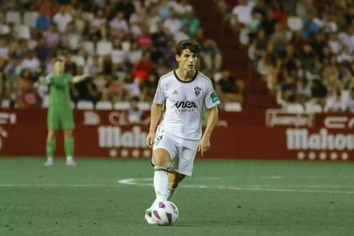 Un jugador de fútbol en el campo, vestido con un uniforme blanco, controla el balón mientras un portero de uniforme verde se encuentra en el fondo.