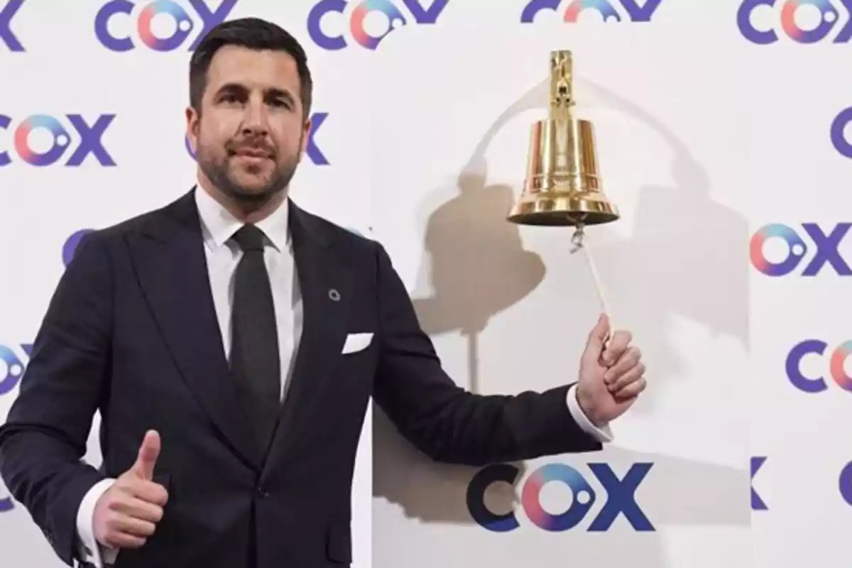 Hombre de traje tocando una campana dorada y haciendo señal de aprobación con el pulgar frente a un fondo con el logo de COX