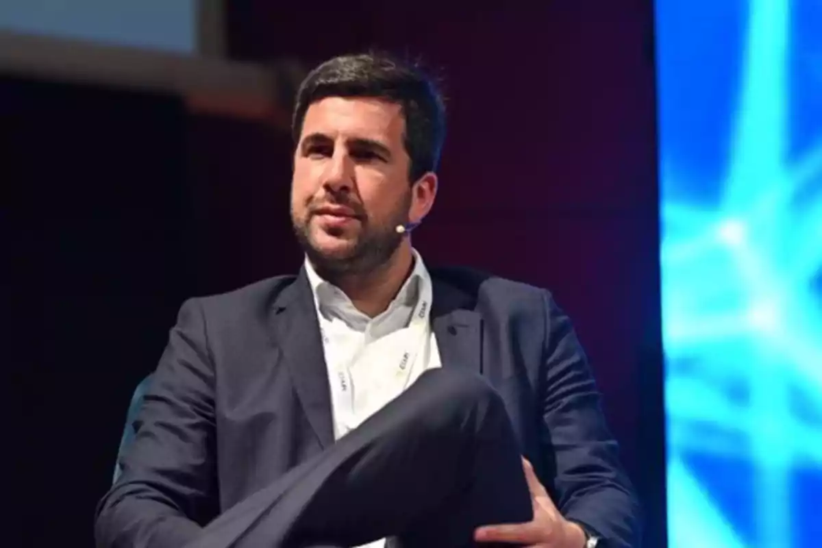 Hombre con barba y cabello oscuro vestido con traje azul marino y camisa blanca sentado en un escenario con fondo azul y micrófono en la mejilla Hombre con barba y cabello oscuro vestido con traje azul marino y camisa blanca sentado en un escenario con fondo azul y micrófono en la mejilla