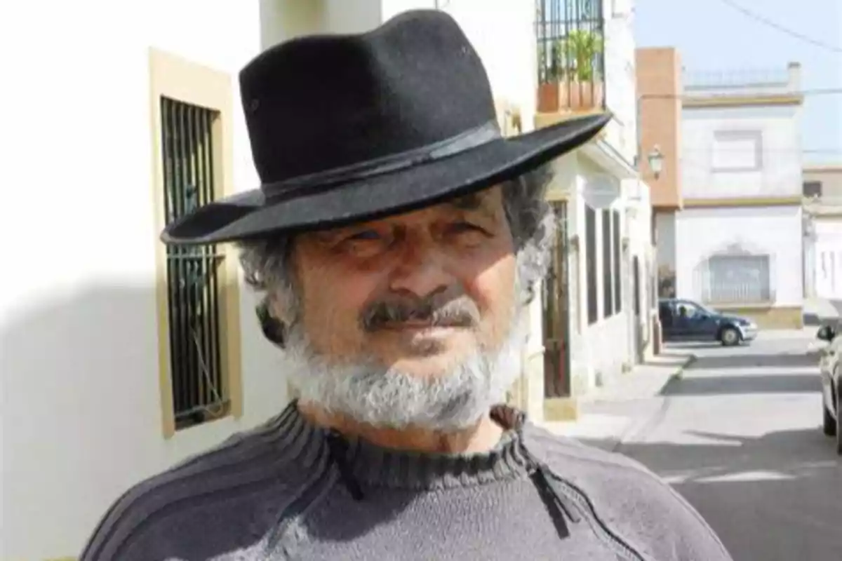 Un hombre con barba y sombrero negro está de pie en una calle soleada.