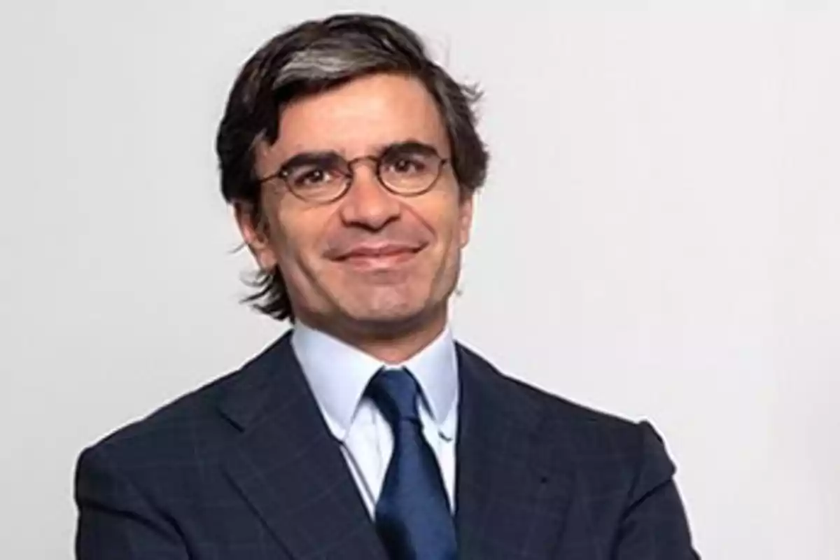 Hombre con gafas y traje oscuro sonriendo frente a un fondo claro.