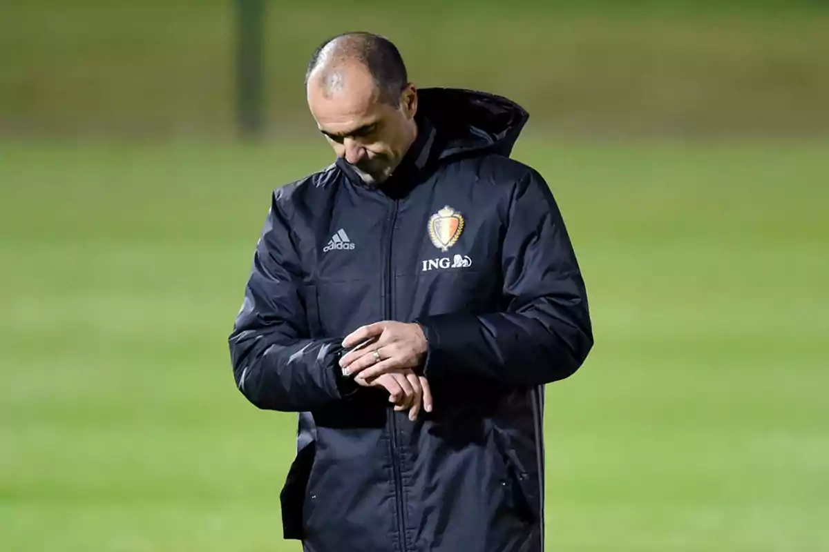 Entrenador de fútbol con chaqueta negra de la selección de Bélgica mirando su reloj en un campo de entrenamiento Entrenador de fútbol con chaqueta negra de la selección de Bélgica mirando su reloj en un campo de entrenamiento
