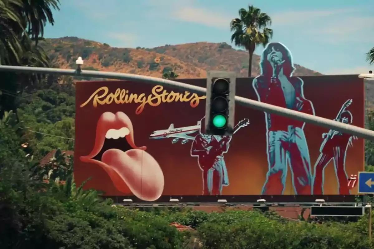 Un cartel publicitario de los Rolling Stones con su icónico logo de lengua y labios, junto a imágenes de los miembros de la banda, se encuentra detrás de un semáforo en verde, con un paisaje montañoso y palmeras al fondo.