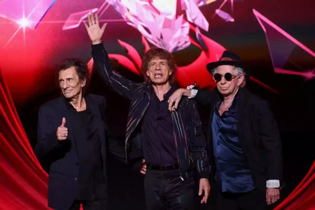 Tres hombres posando en un escenario con un fondo rojo y negro, uno de ellos levantando la mano y otro haciendo un gesto de aprobación con el pulgar.