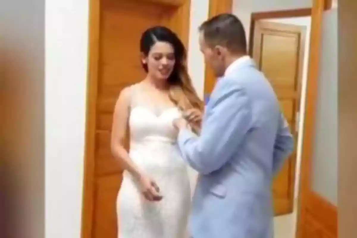 Una pareja vestida elegantemente en un pasillo interior, con la mujer usando un vestido blanco y el hombre un traje azul claro.