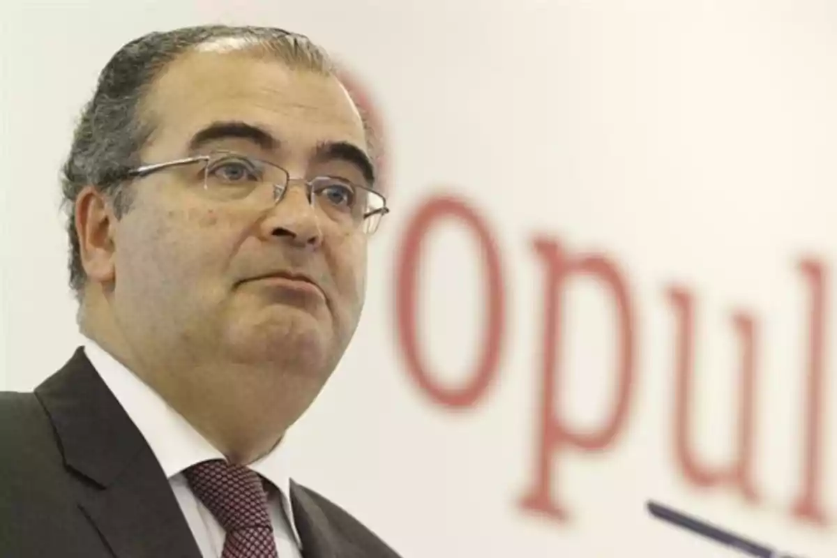 Un hombre con gafas y traje oscuro está de pie frente a un fondo blanco con letras rojas desenfocadas.