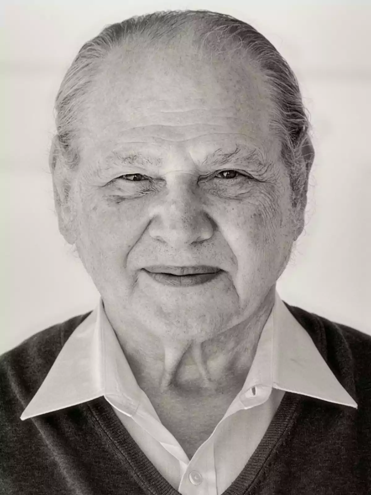 Retrato en blanco y negro de un hombre mayor con cabello canoso y expresión seria, vistiendo una camisa blanca y un suéter oscuro.