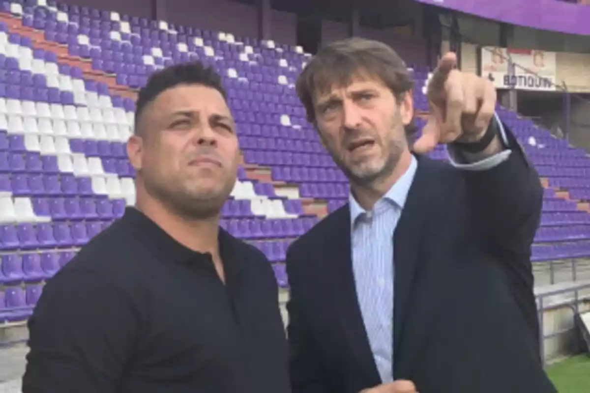 Dos hombres en un estadio con asientos morados y blancos, uno de ellos señalando hacia adelante.