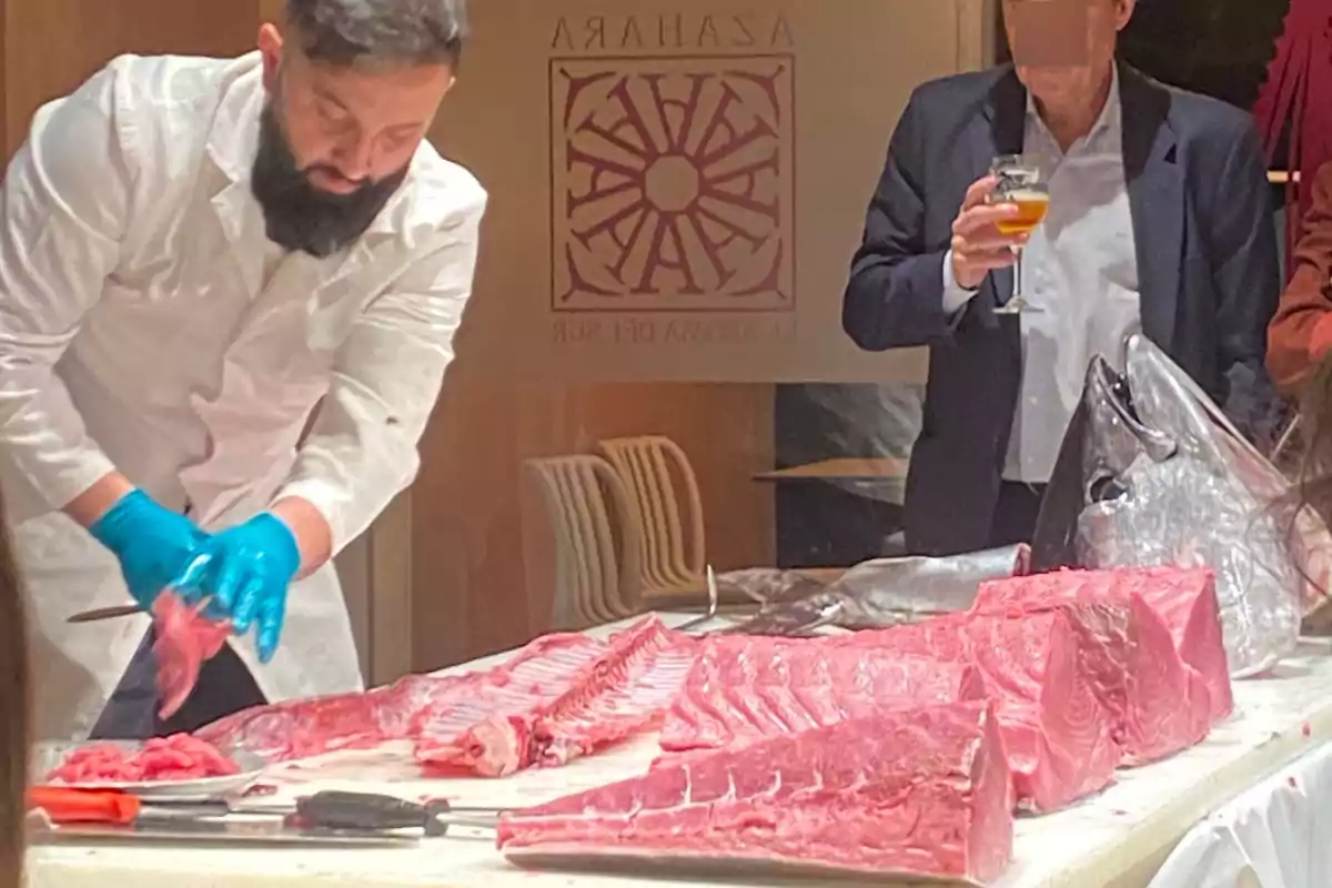 Un chef con guantes azules corta un gran pescado en una mesa mientras un hombre observa con una copa en la mano.