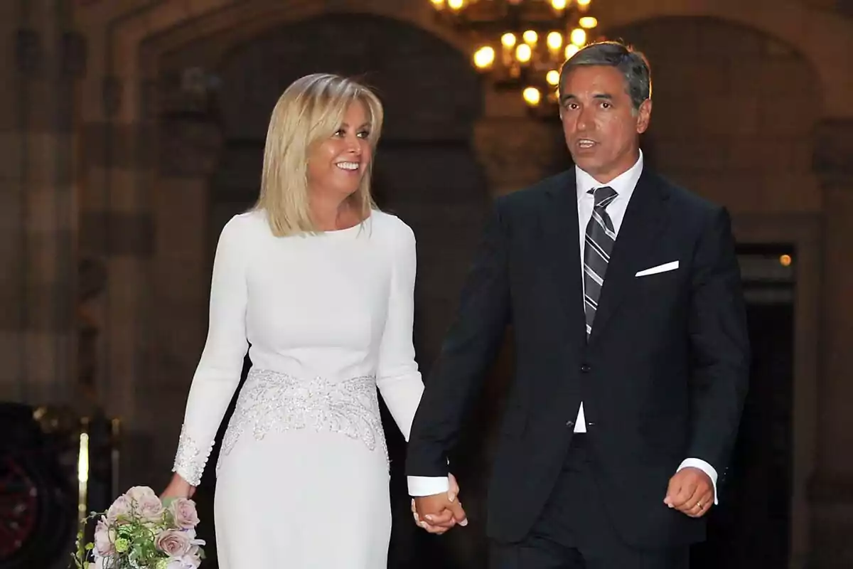Una pareja elegantemente vestida, ella con un vestido blanco y él con un traje oscuro, camina de la mano mientras ella sostiene un ramo de flores.