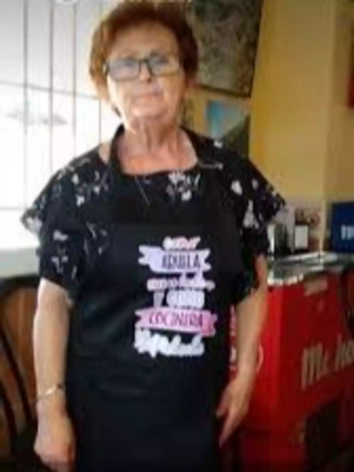 Una mujer mayor con gafas y cabello corto lleva un delantal negro con texto en colores claros, está de pie en un entorno interior con una máquina expendedora roja al fondo. Una mujer mayor con gafas y cabello corto lleva un delantal negro con texto en colores claros, está de pie en un entorno interior con una máquina expendedora roja al fondo.