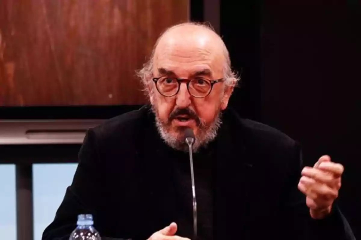 Un hombre con gafas y barba hablando en un micrófono durante un evento en interiores.