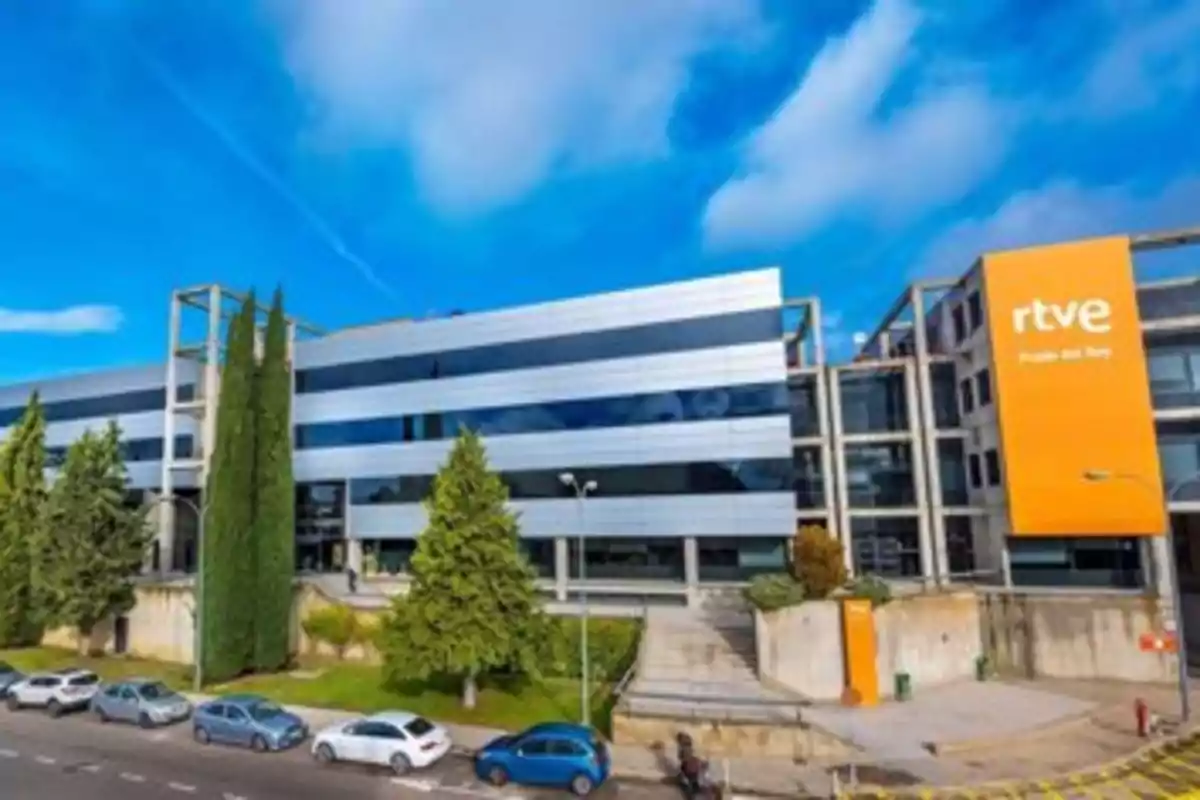 Edificio moderno de RTVE en Prado del Rey con cielo azul y algunos árboles y coches estacionados en la calle.