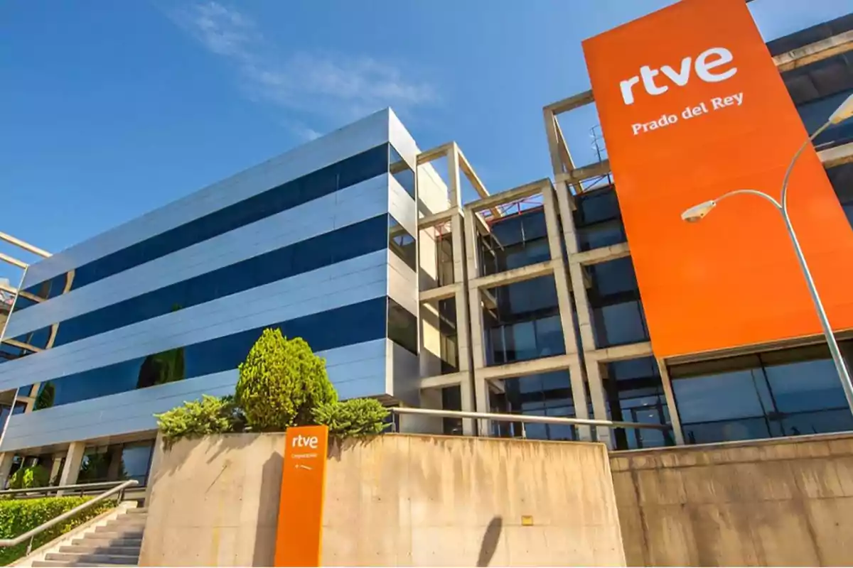 Edificio moderno con fachada de vidrio y estructura de concreto con un cartel naranja que dice RTVE Prado del Rey.