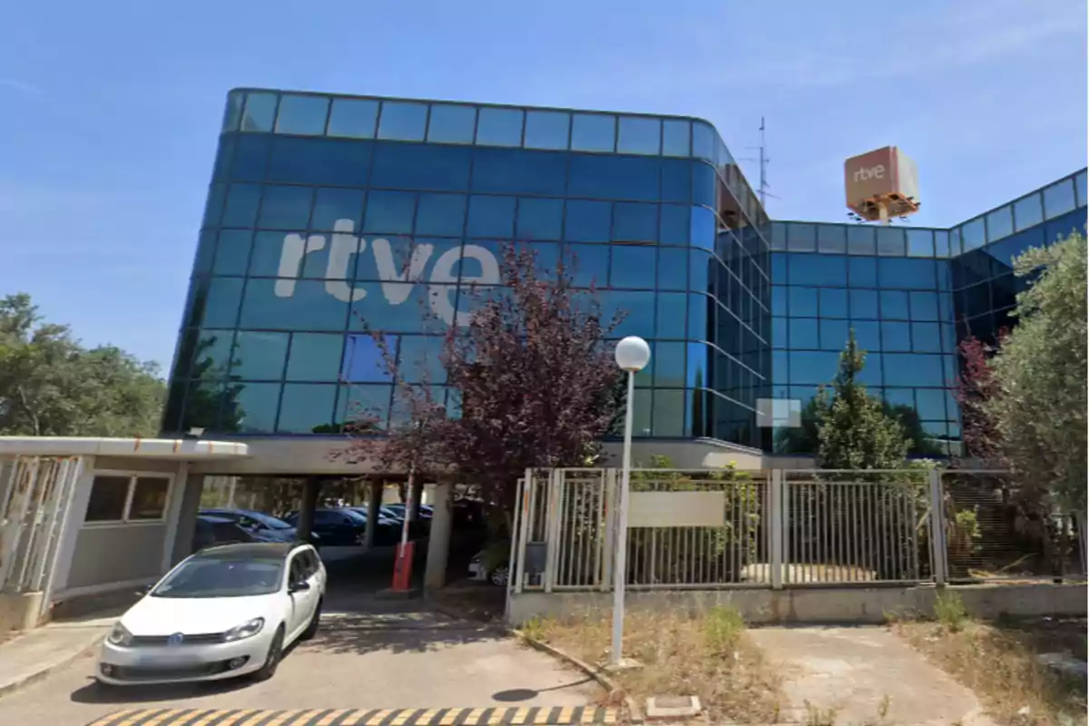 Edificio moderno de cristal azul con el logo de rtve en la fachada y un coche blanco entrando al estacionamiento