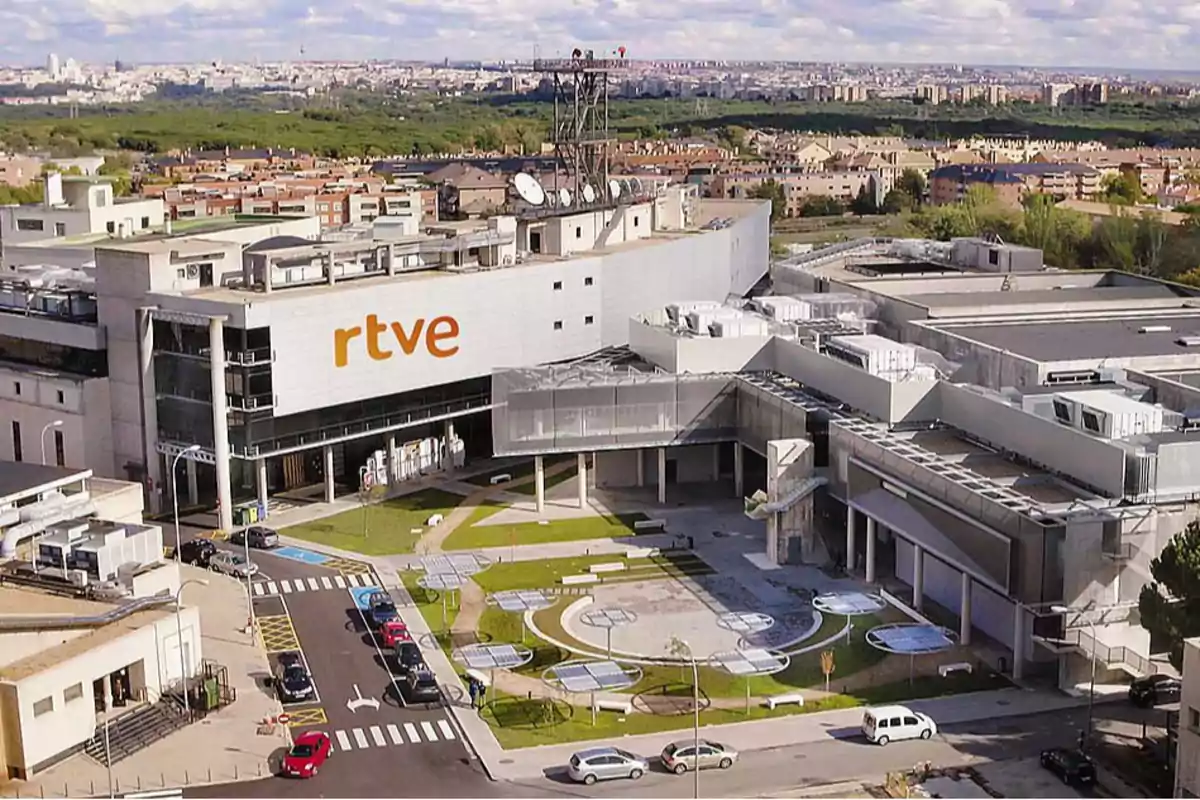 Edificio de RTVE con un amplio patio y estacionamiento, rodeado de áreas verdes y una vista panorámica de la ciudad al fondo.
