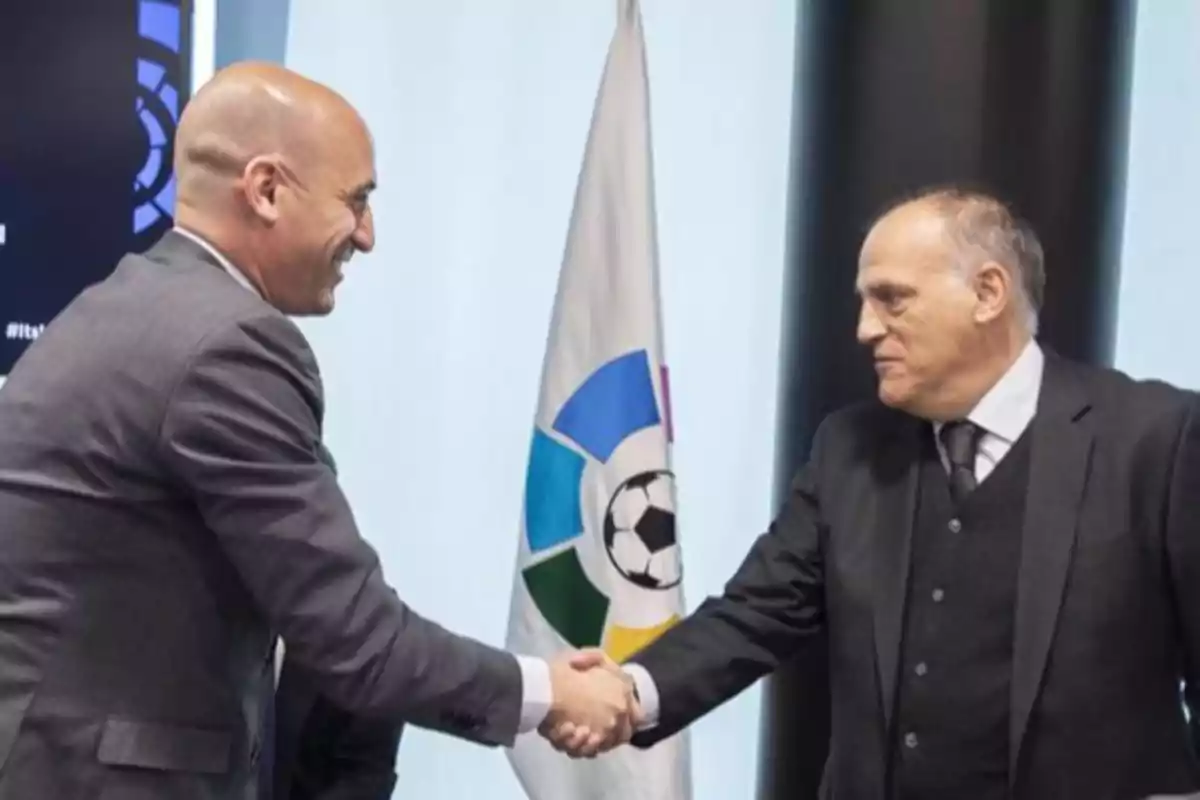 Dos hombres de traje se dan la mano frente a una bandera con un balón de fútbol en el centro.