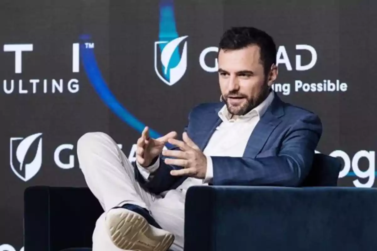Hombre con barba y saco azul sentado en un sillón oscuro gesticulando con las manos durante una conferencia Hombre con barba y saco azul sentado en un sillón oscuro gesticulando con las manos durante una conferencia