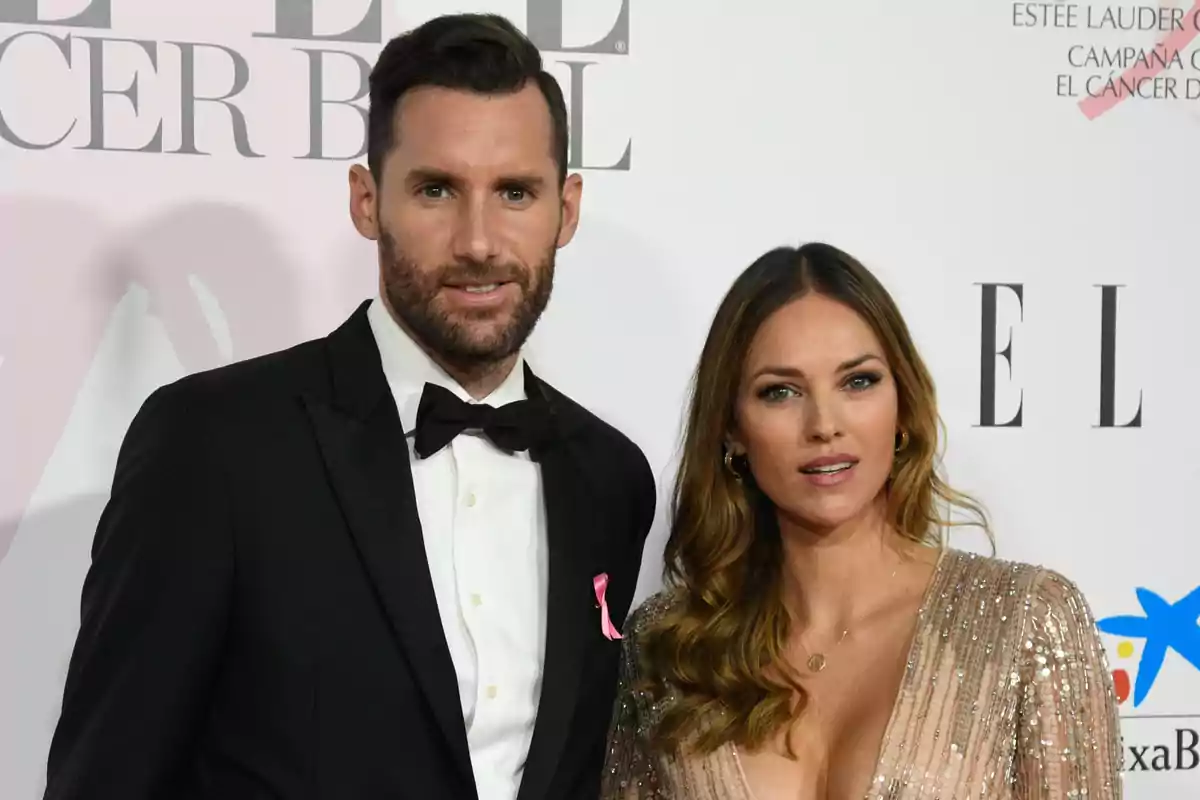 Una pareja elegantemente vestida posa en un evento de gala con un fondo que muestra logotipos de marcas y campañas.