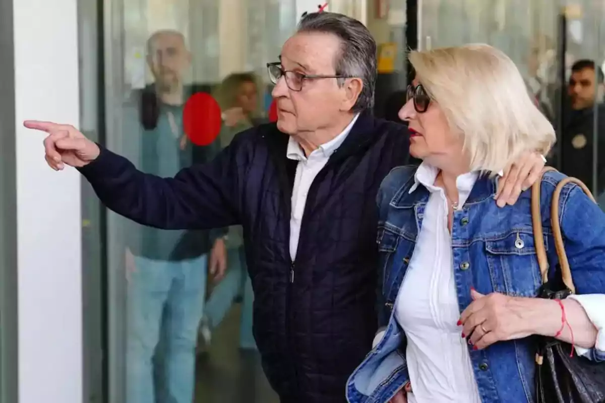 Un hombre y una mujer de mediana edad están de pie juntos, el hombre señala algo a lo lejos mientras la mujer lo observa, ambos llevan chaquetas casuales y gafas de sol.