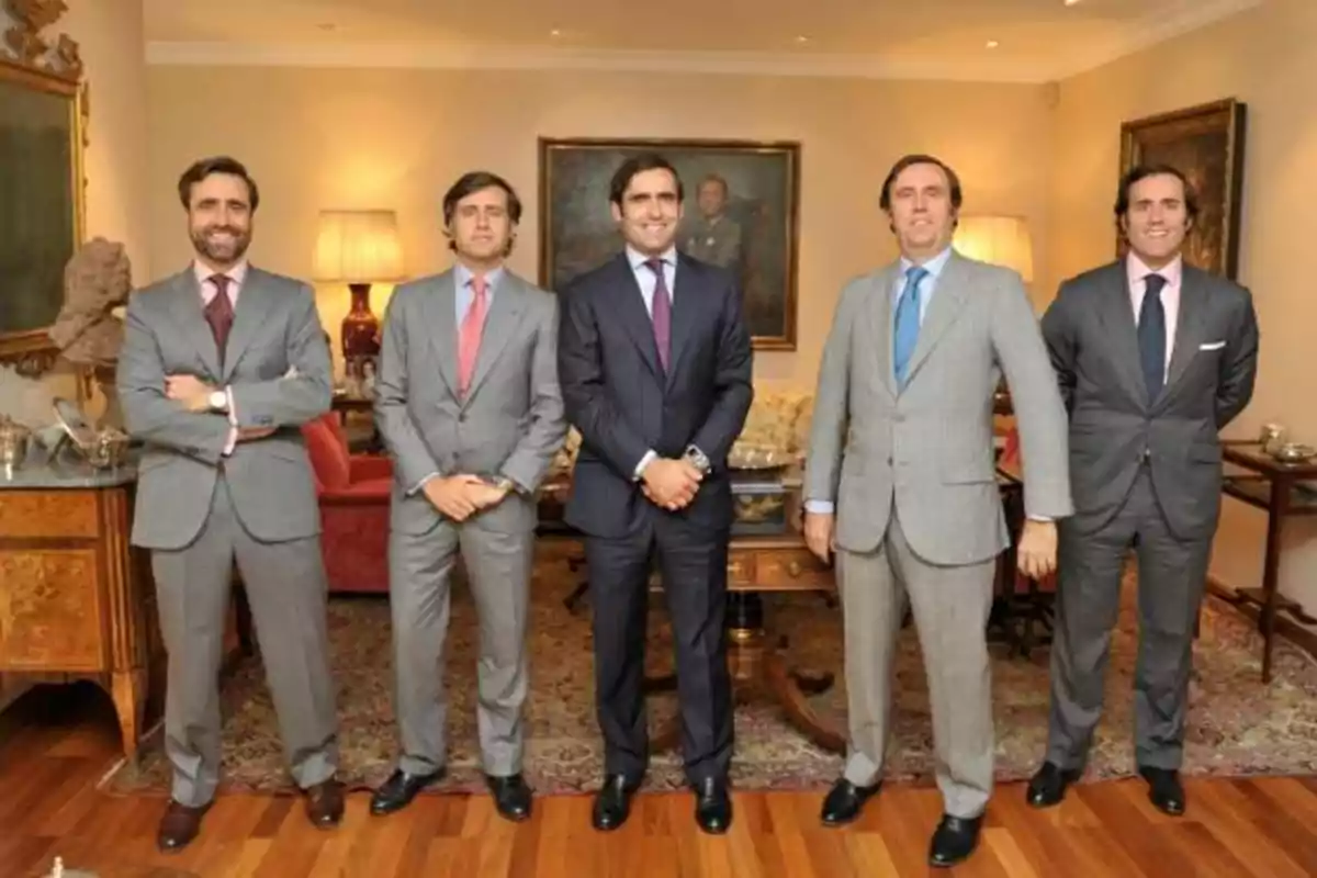 Cinco hombres de traje posan en una sala elegantemente decorada con muebles antiguos y lámparas.