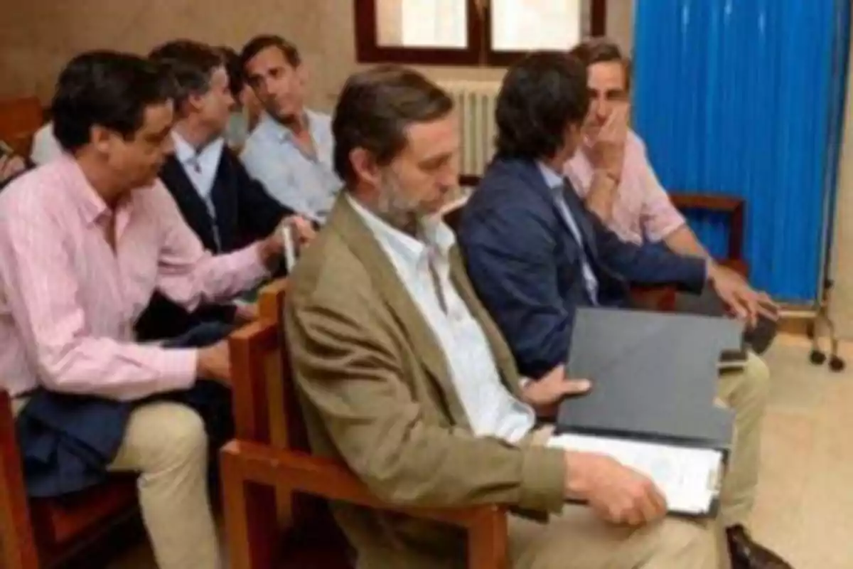 Un grupo de hombres sentados en una sala, algunos conversando entre ellos y otros revisando documentos.