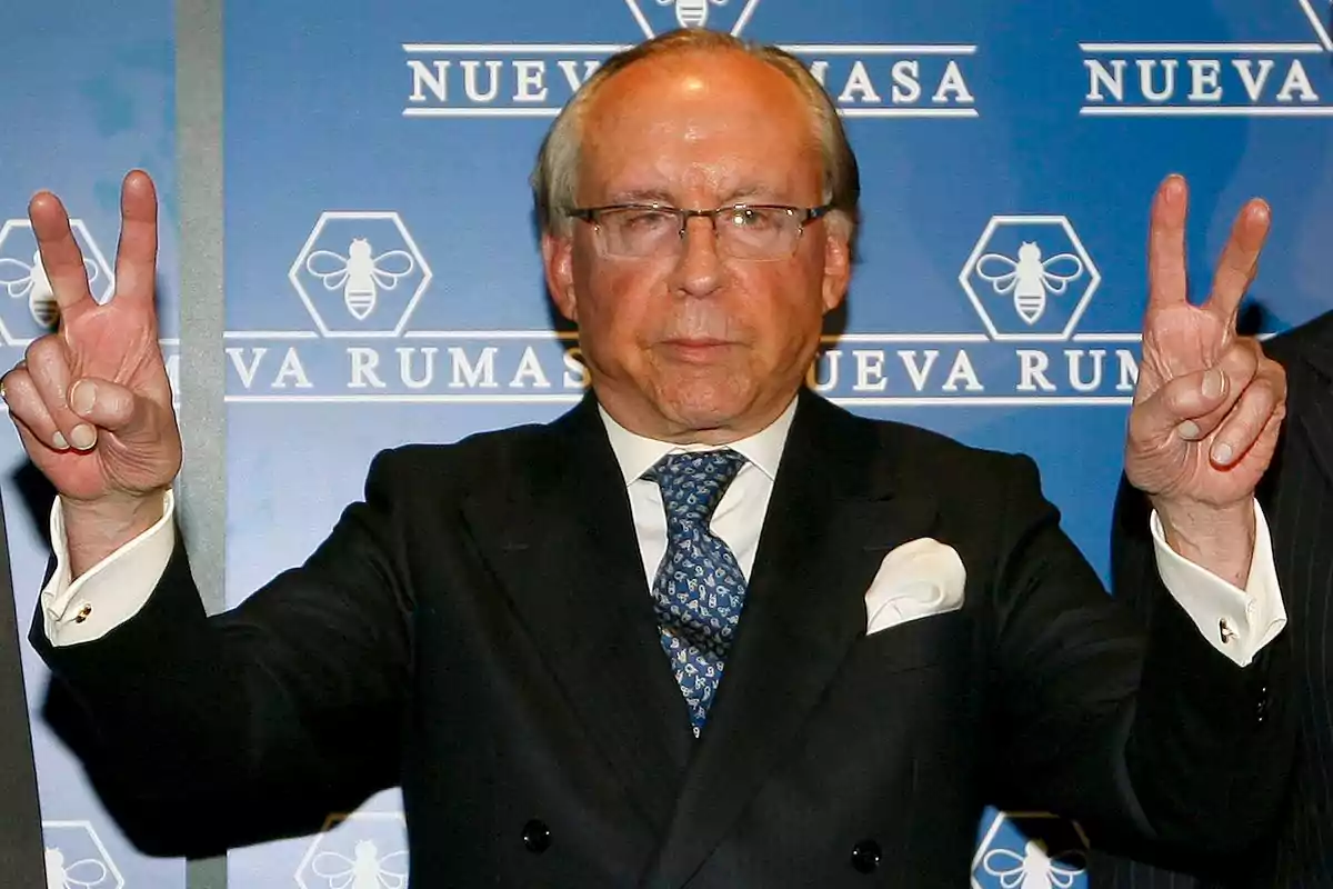 Un hombre mayor con gafas y traje oscuro hace el signo de la victoria con ambas manos frente a un fondo azul que dice "Nueva Rumasa".