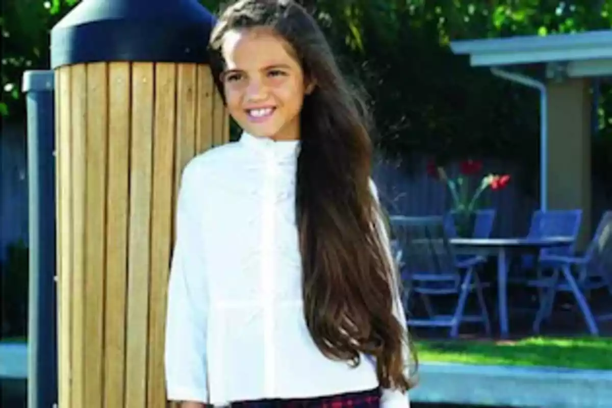 Niña con cabello largo y suelto, vestida con blusa blanca y falda a cuadros, sonriendo al aire libre junto a una estructura de madera.