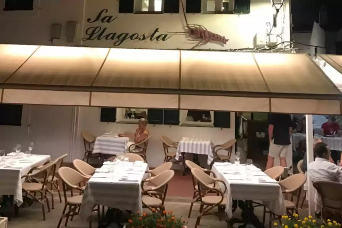 Una terraza de restaurante con mesas y sillas vacías, con una persona sentada en una mesa, bajo un toldo beige y un letrero que dice "La Llagosta".