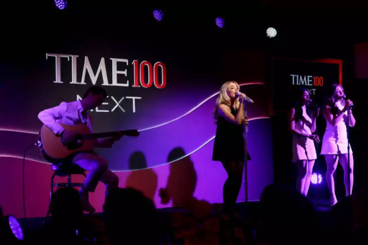 Un grupo musical se presenta en el evento TIME100 Next, con un guitarrista y tres cantantes en el escenario iluminado.