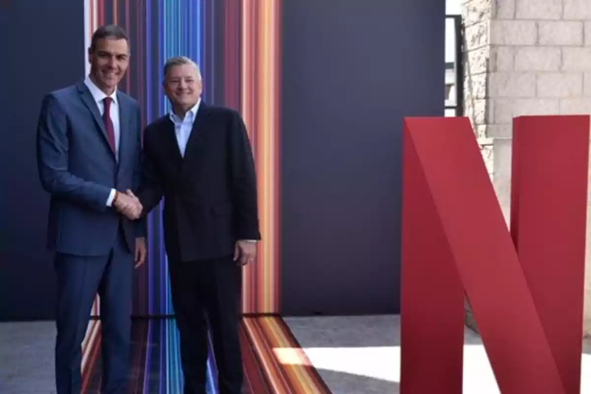 Dos hombres de traje se dan la mano frente a una gran letra N roja y un fondo con líneas de colores verticales.