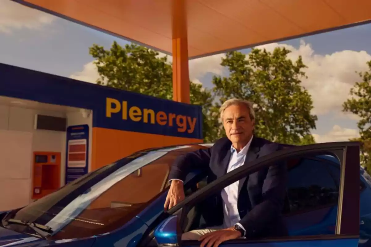 Hombre de cabello canoso con saco oscuro apoyado en la puerta de un auto azul en una estación de servicio llamada Plenergy con árboles y cielo azul al fondo