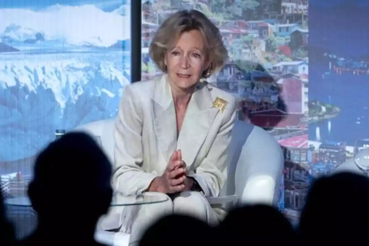 Una mujer sentada en un sillón blanco, vestida con un traje claro, hablando en un evento con un fondo de imágenes de paisajes.