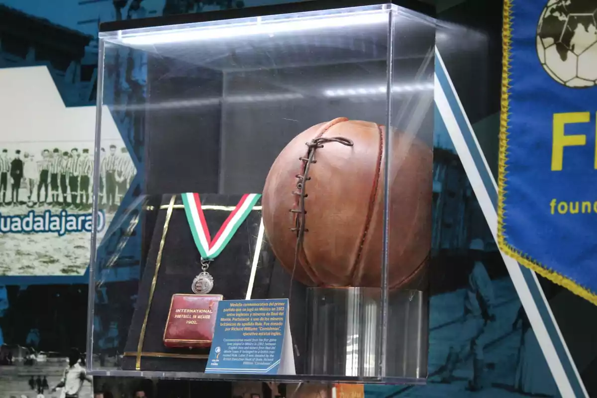 Un balón de fútbol antiguo y una medalla conmemorativa se exhiben en una vitrina iluminada, con un fondo que muestra una imagen histórica de un equipo de fútbol.