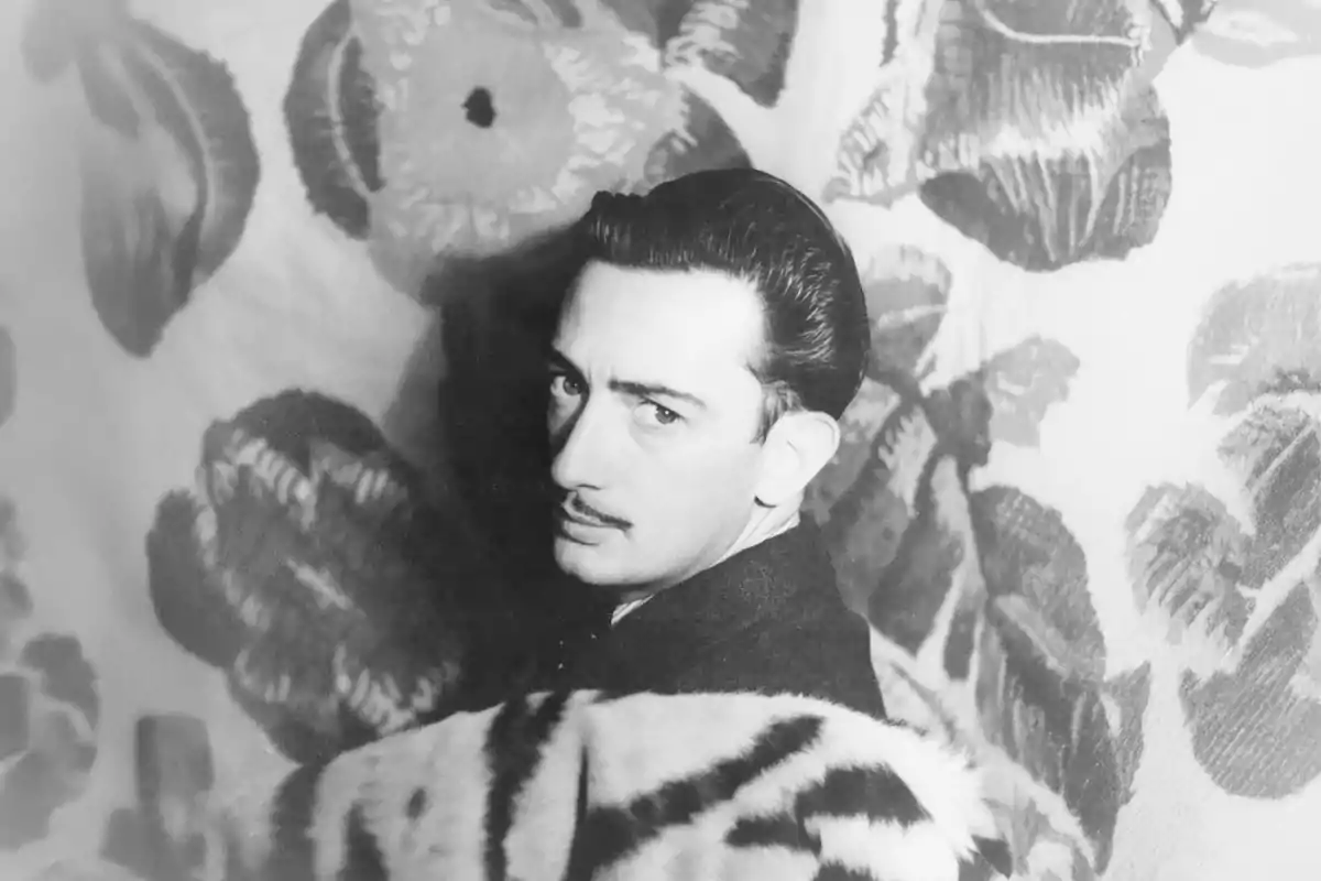 Un hombre con bigote y cabello peinado hacia atrás mira hacia la cámara mientras está envuelto en una tela con estampado de tigre, con un fondo de diseño floral. Un hombre con bigote y cabello peinado hacia atrás mira hacia la cámara mientras está envuelto en una tela con estampado de tigre, con un fondo de diseño floral.